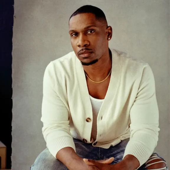 A man wearing a white cardigan, white shirt, and gold chain sits facing the camera with a neutral expression.