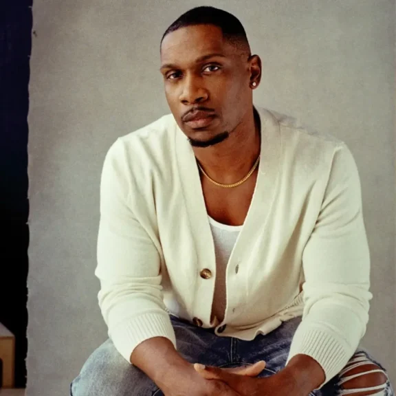 A man wearing a white cardigan, white shirt, and jeans sits on a stool against a neutral background, looking directly at the camera.