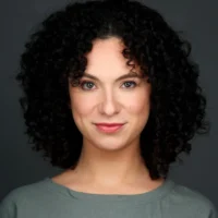 A person with curly dark hair and light skin is facing the camera and smiling slightly, wearing a gray top against a plain dark background.