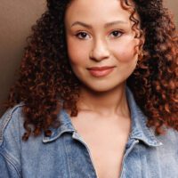 A person with curly hair, wearing a denim jacket and a nose ring, stands in front of a plain brown background, looking directly at the camera.