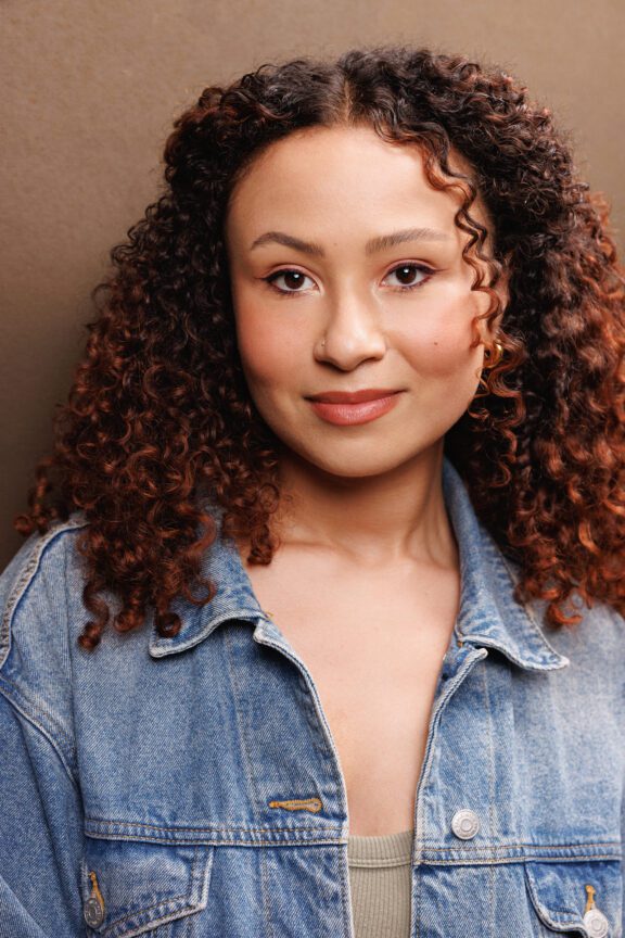 A person with curly hair, wearing a denim jacket and a nose ring, stands in front of a plain brown background, looking directly at the camera.