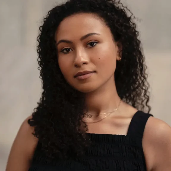 A woman with curly dark hair and medium skin tone wearing a black sleeveless top looks at the camera against a neutral background.