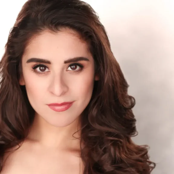 A woman with long, wavy brown hair and brown eyes looks directly at the camera, with a neutral expression and a light background.