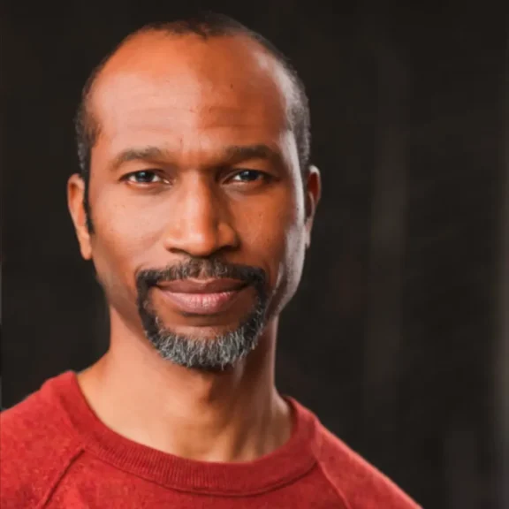 A man with short hair and a salt-and-pepper goatee wears a red sweater and looks at the camera against a dark background.