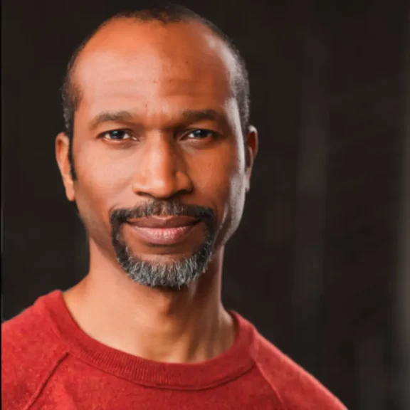 A man with short hair and a graying beard wears a red sweater and stands in front of a dark background, looking at the camera.