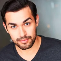 A man with dark hair and a beard looks at the camera, wearing a dark gray V-neck shirt against a blurred background.