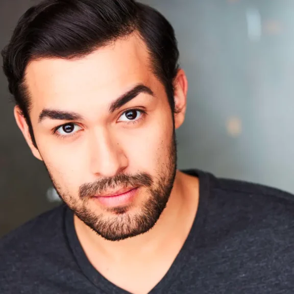 A man with dark hair and a beard looks at the camera, wearing a dark gray V-neck shirt against a blurred background.