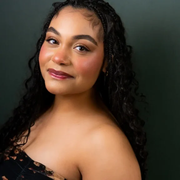A woman with long curly hair, wearing a strapless top, looks at the camera against a dark green background.