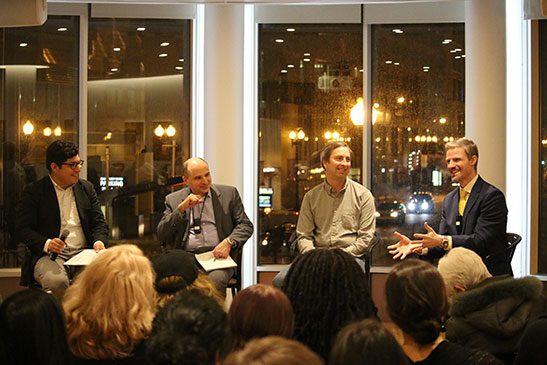 A group of panelists sit in front of an audience at an evening event. The background behind the panelists are large windows.