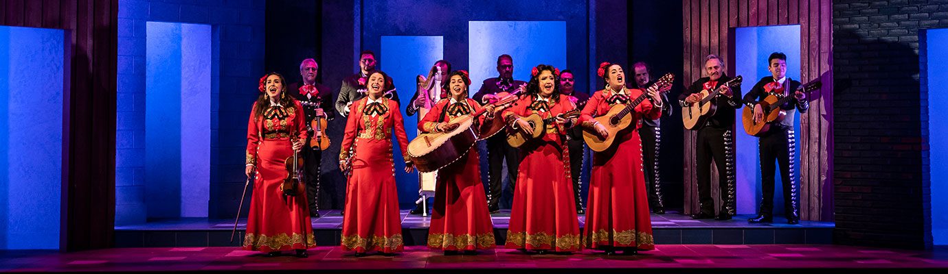 Two rows of actors on stage performing mariachi music. In the front row, female musicians are dressed in bright red mariachi costumes and play various instruments. In the back row, male performers are dressed in black mariachi suits and play instruments.