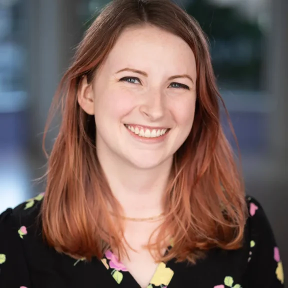 A young woman with shoulder-length auburn hair smiles at the camera. She is wearing a black top with a colorful floral pattern. The background is softly blurred.