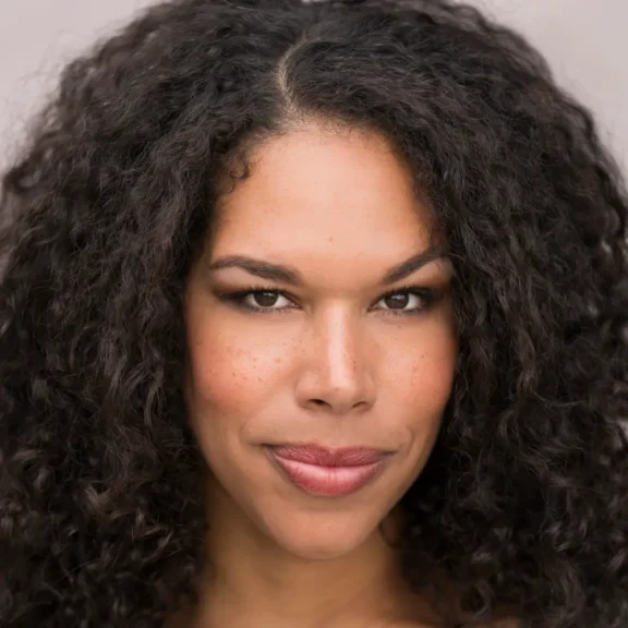 A woman with medium skin tone and curly dark hair looks directly at the camera with a neutral expression.