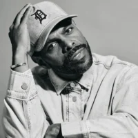 Portrait of a man wearing a baseball cap with a “D” logo and a denim shirt, looking toward the camera with his hand raised to his head.