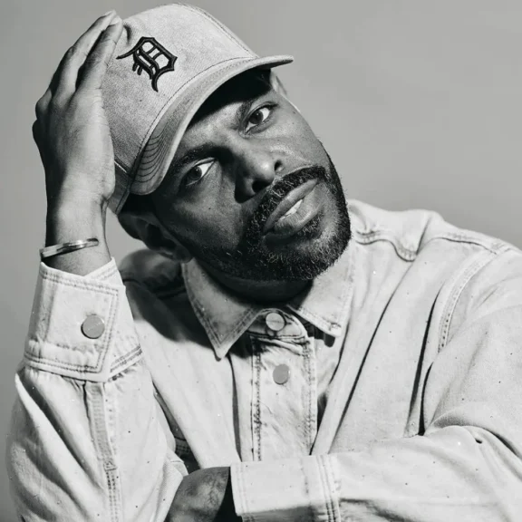 Portrait of a man wearing a baseball cap with a “D” logo and a denim shirt, looking toward the camera with his hand raised to his head.