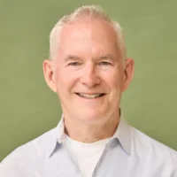 An older man with short white hair wearing a light-colored collared shirt smiles in front of a plain green background.