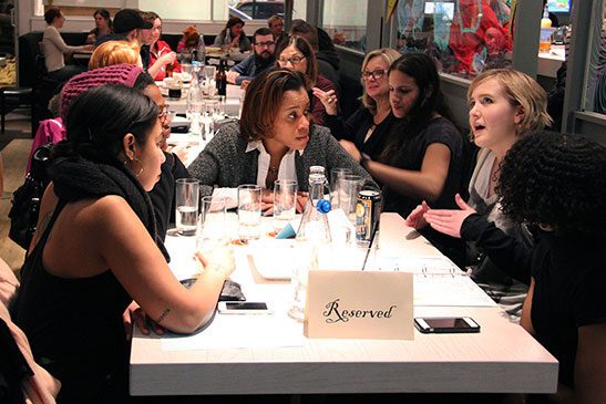 A group of people gather around a table having a conversation. There is a reserved sign at the head of the table.