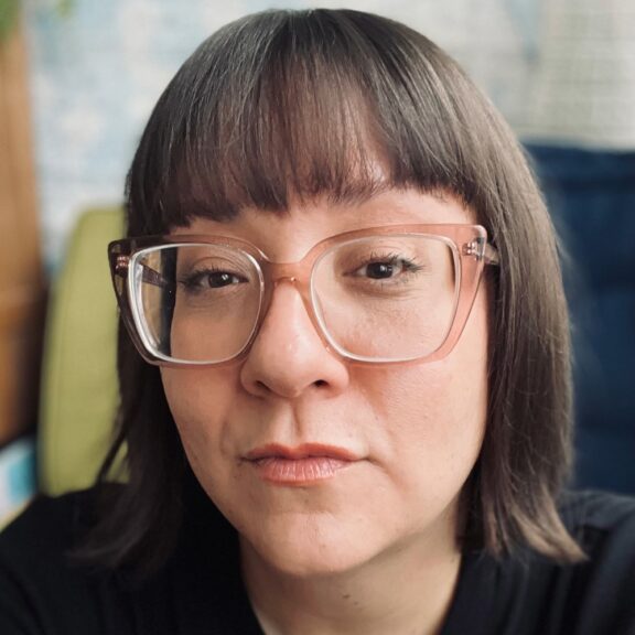 A woman with straight brown hair, blunt bangs, and large clear-framed glasses looks directly at the camera with a neutral expression.