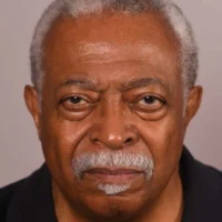 An older man with short gray hair and a mustache looks directly at the camera with a neutral expression. He is wearing a dark shirt.