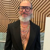 A man with a gray beard and glasses wears a black blazer with no shirt, layered necklaces, and stands in front of a geometric wooden wall.
