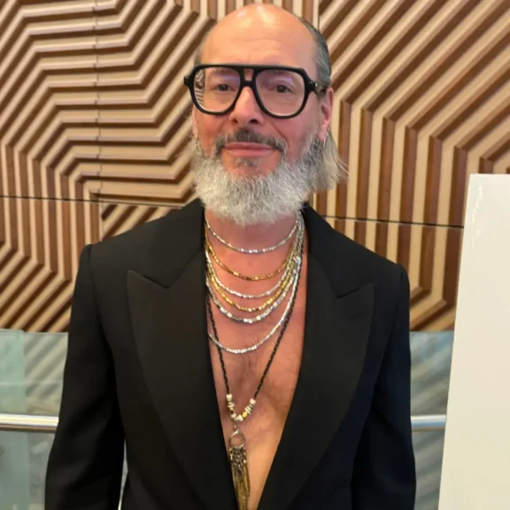 A man with a gray beard and glasses wears a black blazer with no shirt, layered necklaces, and stands in front of a geometric wooden wall.