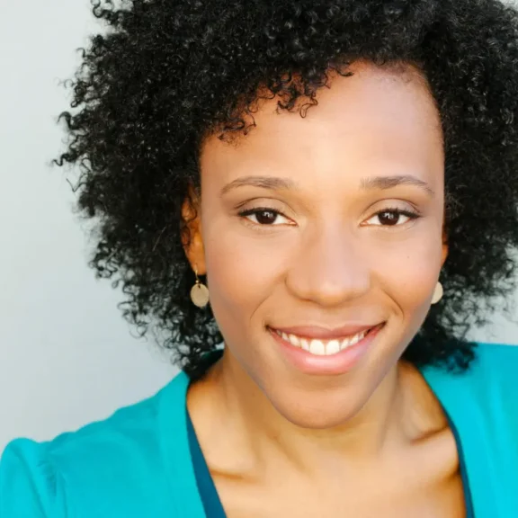 A woman with curly hair smiles at the camera, wearing a teal top and round earrings, posed against a light background.