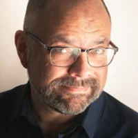 A middle-aged man with glasses, short hair, and a beard is looking at the camera with a slight smile, wearing a dark shirt against a plain light background.