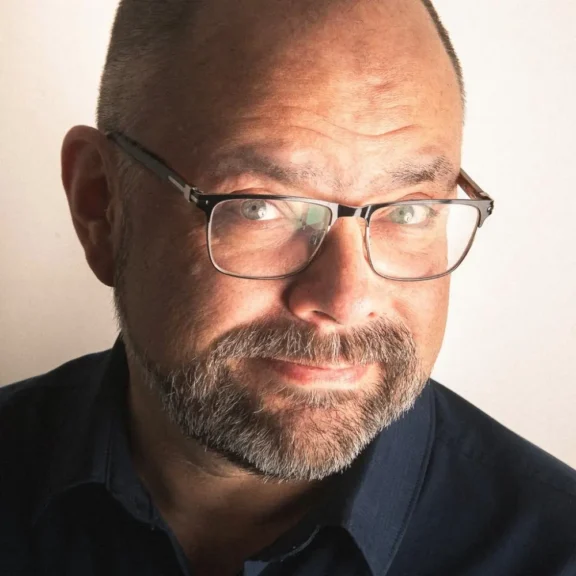 A middle-aged man with glasses, short hair, and a beard is looking at the camera with a slight smile, wearing a dark shirt against a plain light background.