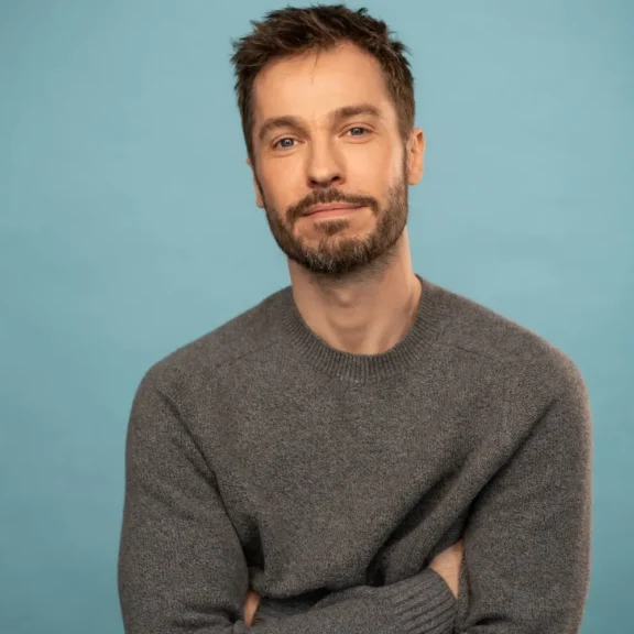 A man with a beard wearing a grey crew-neck sweater, posing with his arms crossed against a blue background.