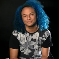 A person with bright blue curly hair wears a black and gray floral-patterned shirt, sitting against a dark background and looking at the camera.