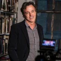 A man in a blazer and gray shirt stands indoors near bookshelves, smiling, with a camera in the foreground.