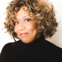 A woman with curly, highlighted hair wearing a black turtleneck sweater, looking at the camera with a slight smile against a light background.