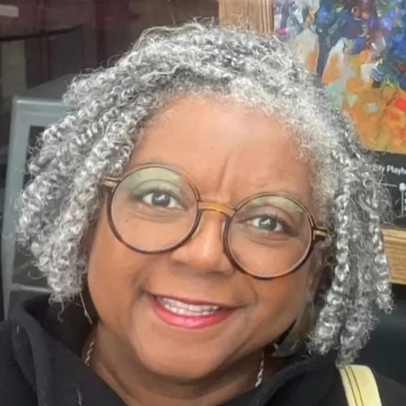 A woman with short, curly gray hair and round glasses smiles at the camera. She is wearing a black top and is positioned in front of a colorful painting.