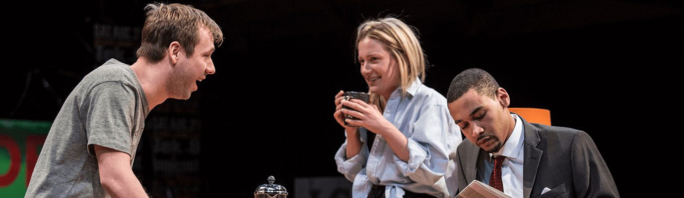 Three actors are captured mid-scene. One actor on the left is excitedly looking at the other two actors. The actor in the middle smiles and holds a coffee mug. The other actor to the right of the image reads a document.