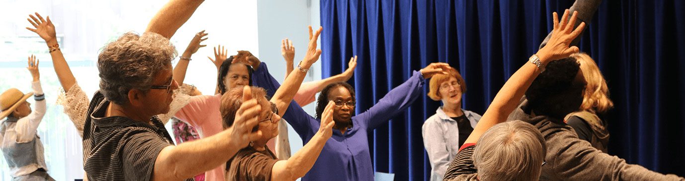 A group of community members in action with their hands raised to the ceiling as they stand in font of a blue curtain.