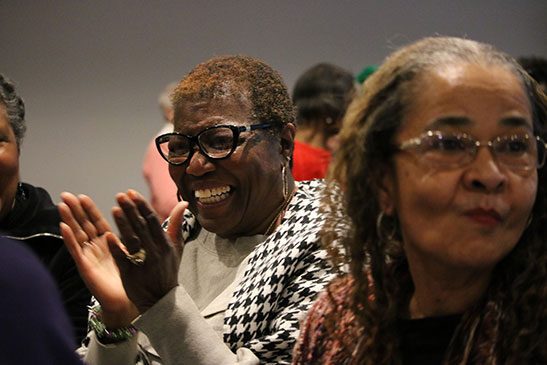 While at an event, one woman who wears glasses is smiling and about to clap her hands while another woman is looking at something to the right of the screen.