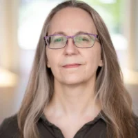 A middle-aged woman with long gray hair and glasses, wearing a dark shirt, stands indoors with a neutral expression.