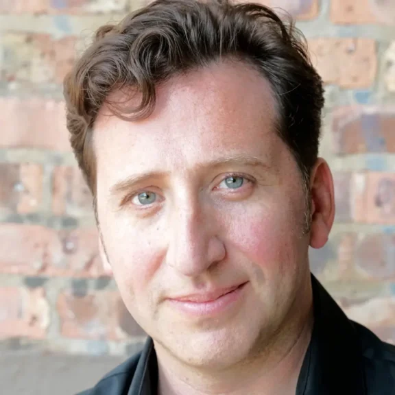 A man with wavy brown hair and blue eyes is looking at the camera, standing in front of a brick wall.