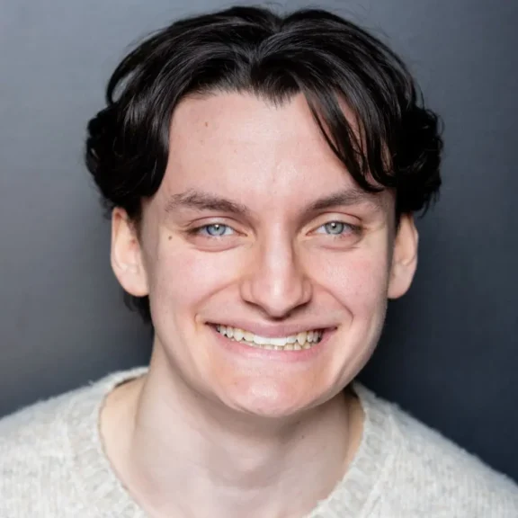 Headshot of a person with dark, wavy hair and light eyes smiling at the camera.
