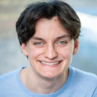 A person with short dark hair and blue eyes is smiling at the camera, wearing a light blue shirt. The background is softly blurred.