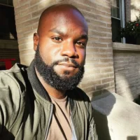 A man with a beard and shaved head sits outside in sunlight, wearing a green jacket and brown shirt, with a brick wall and window in the background.