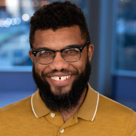 A man with short curly hair, a full beard, and glasses, wearing a mustard yellow polo shirt, smiles at the camera with a blurred cityscape background.