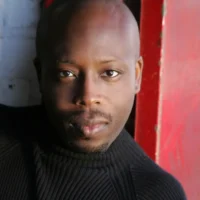 A man with a shaved head wearing a black ribbed turtleneck sweater poses in front of a red and white background.