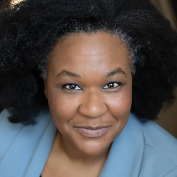 A woman with natural curly black hair and subtle makeup, wearing a light blue blazer, looks directly at the camera with a slight smile.