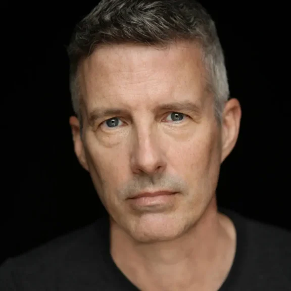 A middle-aged man with short gray hair and blue eyes stares directly at the camera against a black background. He is wearing a dark shirt and has a neutral expression.