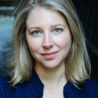 A woman with shoulder-length blonde hair and blue eyes wears a dark blue top and smiles slightly at the camera. The background is blurred and dark.