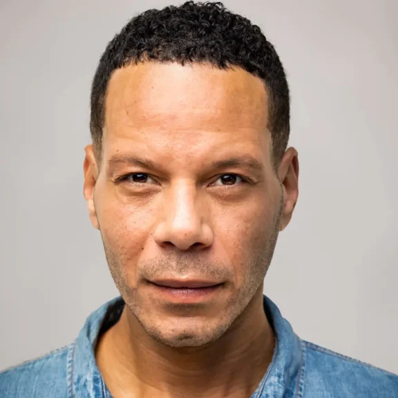 A man with short curly hair and a slight beard is wearing a blue denim shirt and looking directly at the camera against a plain background.