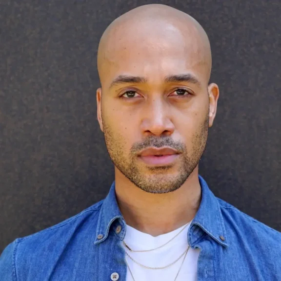 A man with a shaved head and short beard wears a denim shirt over a white t-shirt, standing against a dark background, looking directly at the camera.
