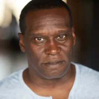 A man with short hair wearing a light gray shirt looks directly at the camera with a neutral expression.