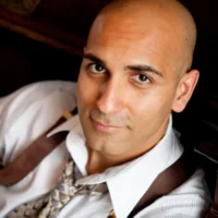Headshot of a man with a shaved head, wearing a white collared shirt, patterned tie, and brown suspenders.