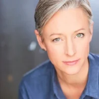 A person with short gray hair and blue eyes, wearing a blue collared shirt, looks at the camera against a blurred dark background.